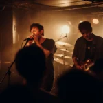 A rock band performing energetically on a small, dimly lit stage in a crowded club.