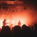 A rock band silhouetted on stage, performing under dramatic pink, orange, and white lights in a dark music venue.