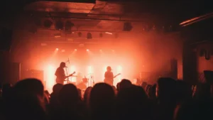 A rock band silhouetted on stage, performing under dramatic pink, orange, and white lights in a dark music venue.