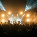 Silhouetted rock band performing on a smoke-filled concert stage with dramatic beams of light before a large crowd