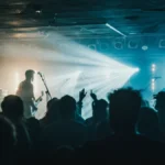 An alternative rock band performs on a small stage with dramatic lighting in front of a silhouetted crowd.