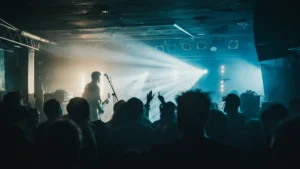 An alternative rock band performs on a small stage with dramatic lighting in front of a silhouetted crowd.