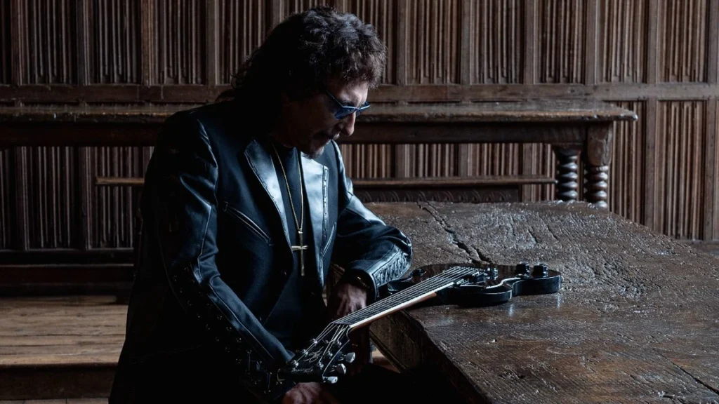 Tony Iommi seated at a long oak table with his black SG guitar in a wood-panelled hall