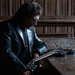 Tony Iommi seated at a long oak table with his black SG guitar in a wood-panelled hall